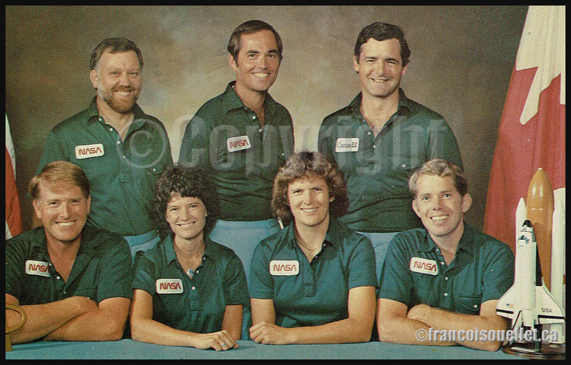 L'astronaute canadien Marc Garneau (rangée du haut, extrême droite) et les astronautes américains choisis par la Nasa pour la mission 41-G avec la navette spatiale Challenger (de gauche à droite et de bas en haut: Jon A.McBride, Sally K. Ride, Kathryn D. Sullivan, David C. Leestma, Paul D.Scully-Power, Robert L. Crippen, Marc Garneau) sur carte postale aviation.
